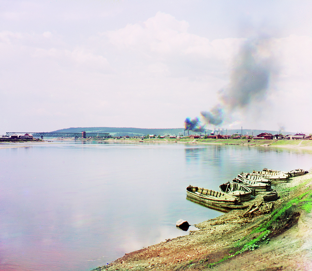 a trichromy by Prokudin-Gorsky showing a few boats in water, a bridge and factory with smoking chimneys in the distance.
