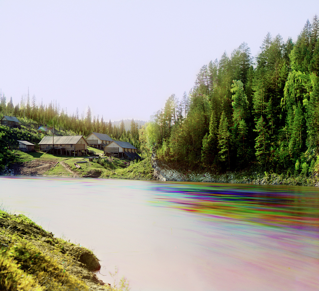 a trichromy by Prokudin-Gorsky showing a fast flowing river exhibiting color ghosting, and a few houses in the distance.