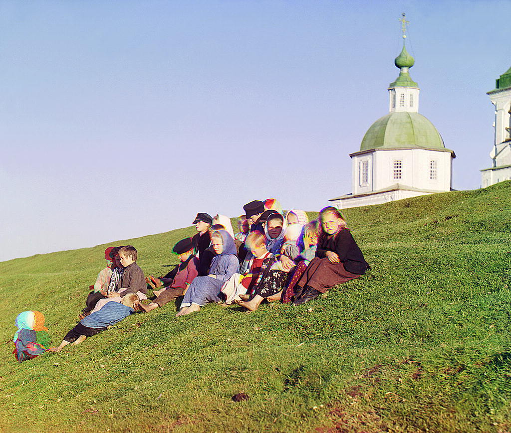 a trichromy by Prokudin-Gorsky showing a group of kids sitting in the sloped grass, a church in the background.